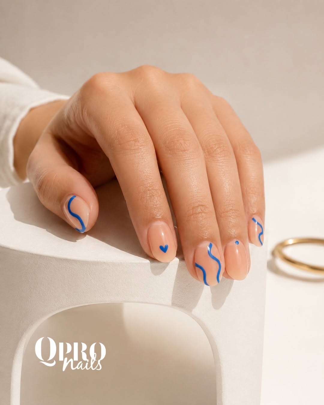 Short natural nails with fine blue line art and a small heart on a white sculptural plinth