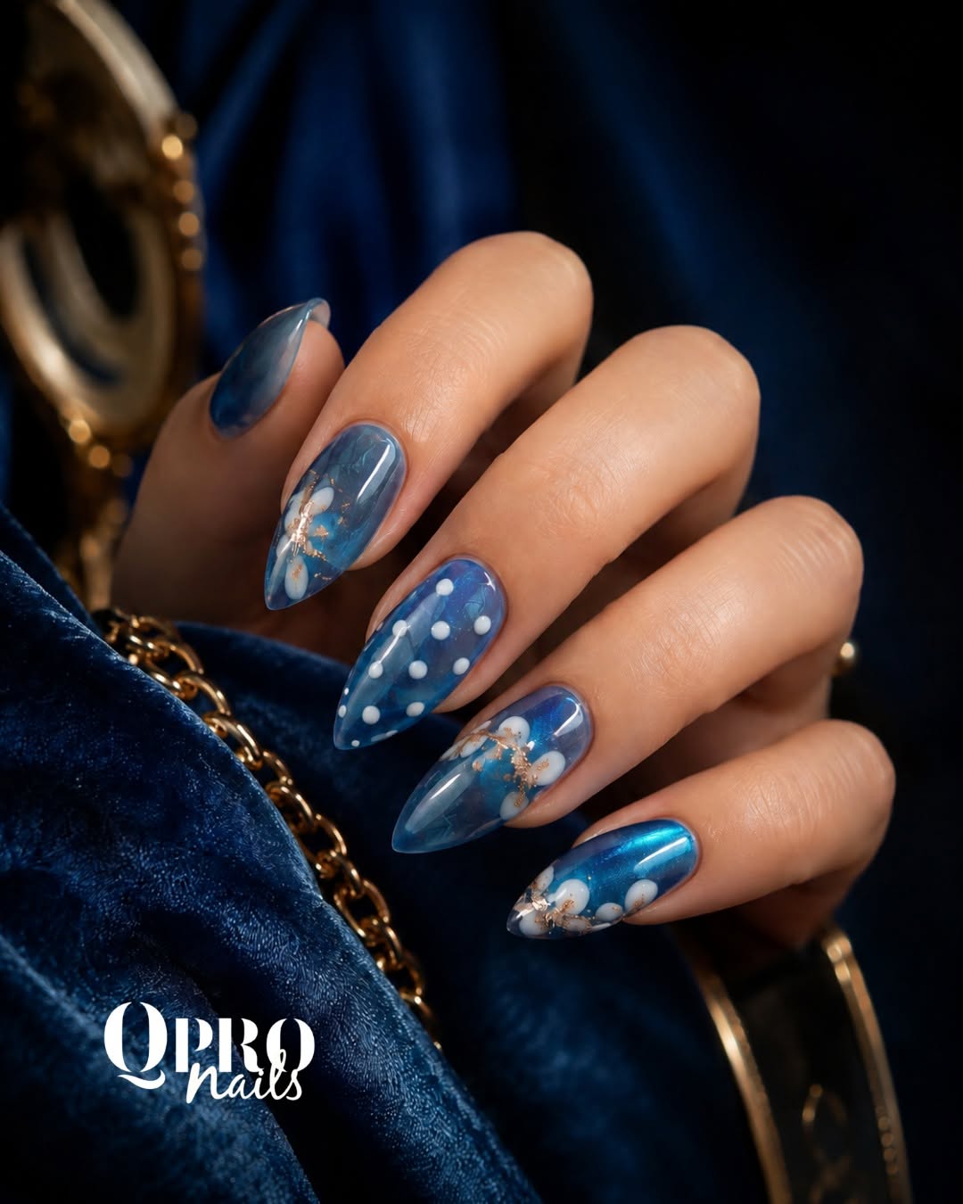 Translucent blue almond nails with white flowers, gold flecks and polka dots against navy velvet